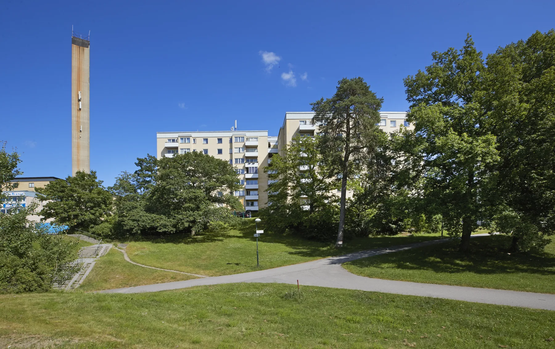 En hög, smal skorsten står bredvid flervåningshus, omgiven av gröna träd och en gräsbevuxen park med korsande asfalterade stigar under en klarblå himmel.