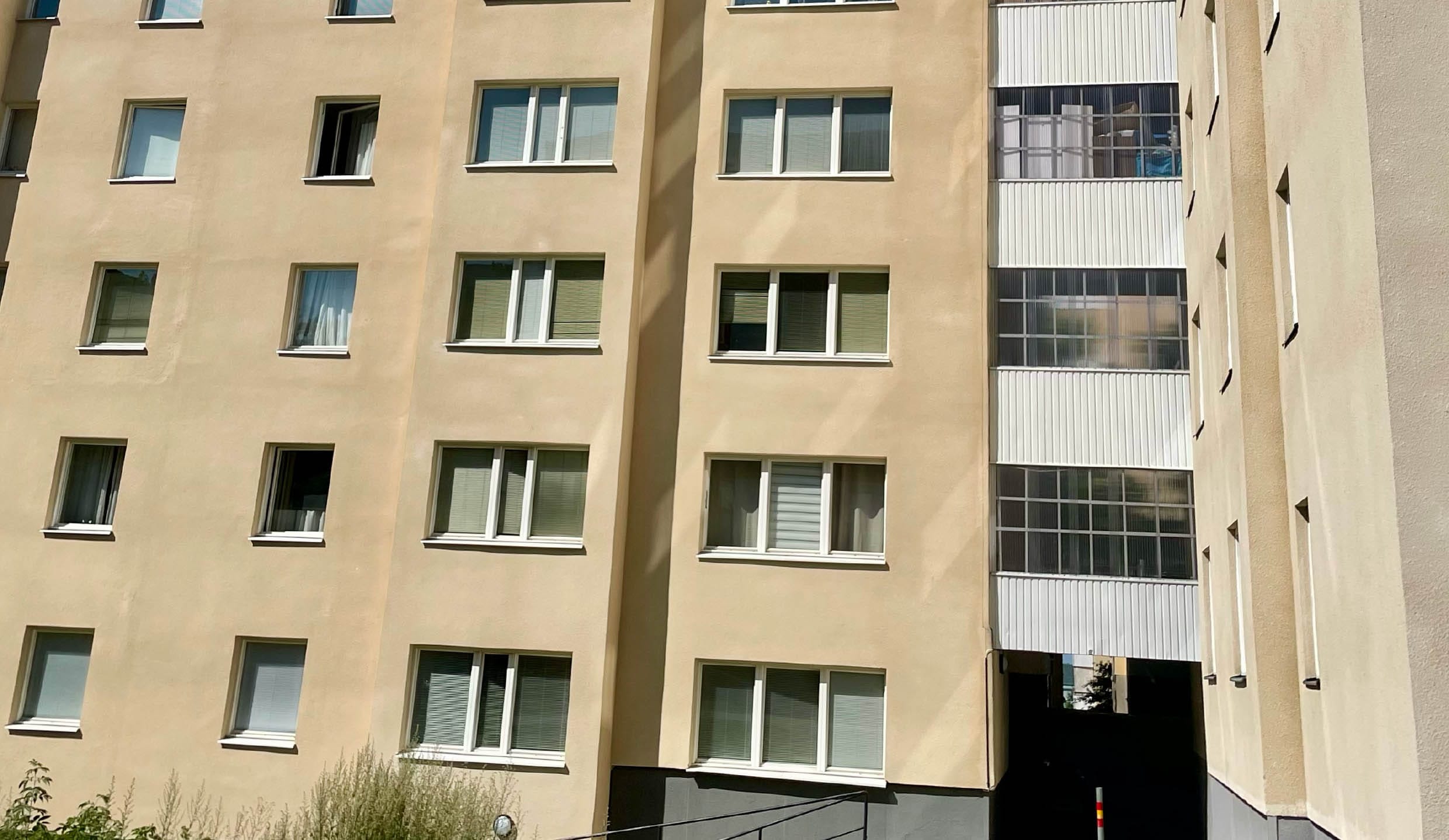 Ett beige flerbostadshus med flera identiska fönster, varav vissa är öppna och andra har persienner. På höger sida finns en täckt gång och längst ner syns lite grönska.